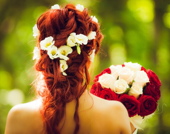 Fotografia przedstawia zbliżenie na tył głowy kobiety. Ma rude, ufryzurowane włosy. W nie wpięte białe kwiaty. Trzyma bukiet.