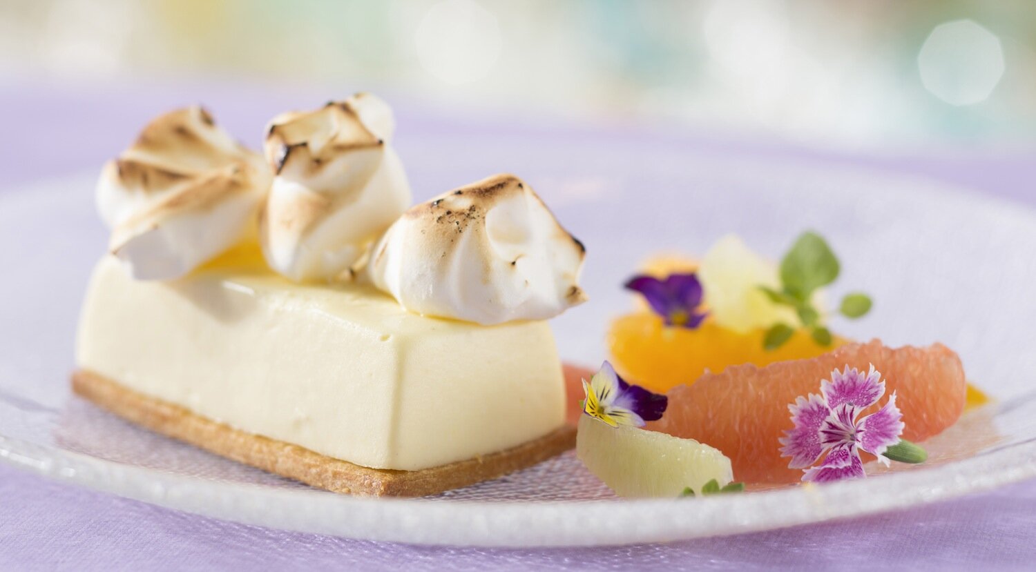 Na zdjęciu widoczny jest deser: ciastko z przypalanymi piankami. Całość ozdobiona jest kolorowymi kwiatami i kawałkiem grejfruta.