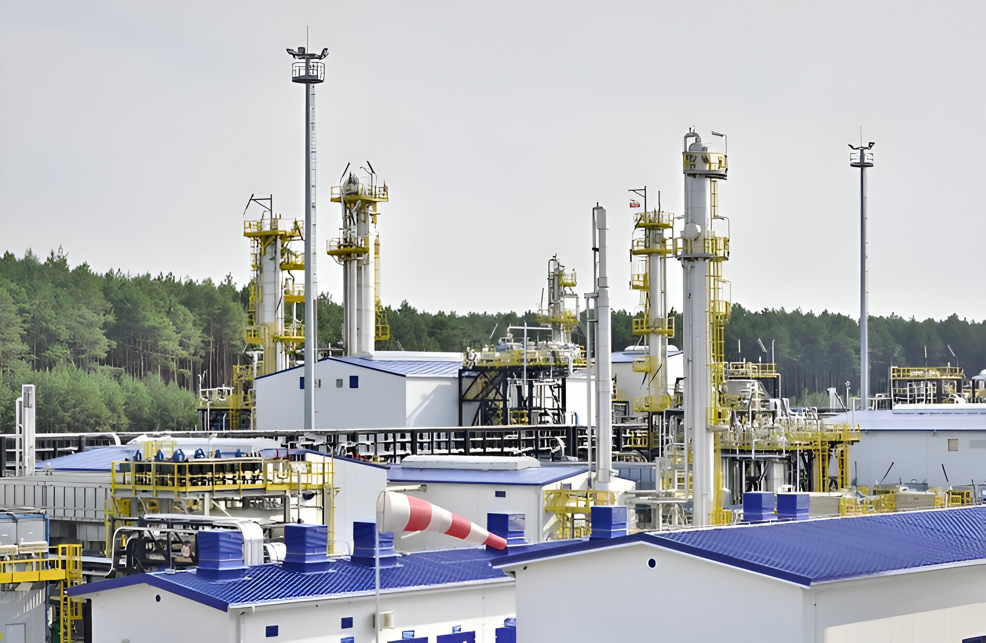 Zdjęcie przedstawia kopalnię ropy naftowej i gazu ziemnego. W tle widać las i jasnoszare niebo. Na pierwszym planie znajdują się białe budynki o niebieskich dachach. Wśród budynków stoi kilka wysokich kominów.