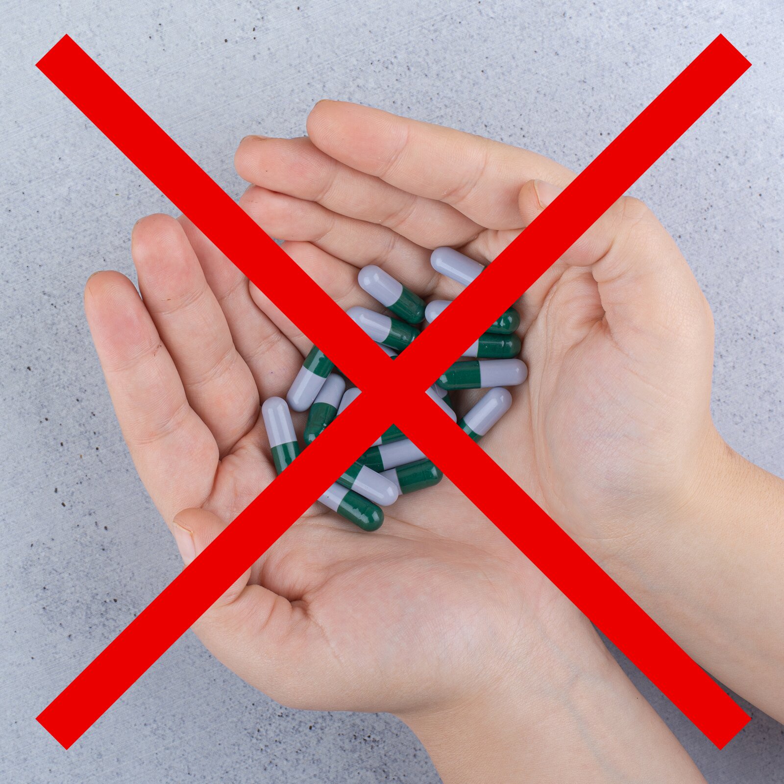 Zdjęcie przedstawia dwie złożone obok siebie dłonie, które są wypełnione owalnymi szaro‑zielonymi tabletkami. Na ilustrację nałożony jest czerwony znak iks, który przekreśla zdjęcie i symbolizuje zakaz.  