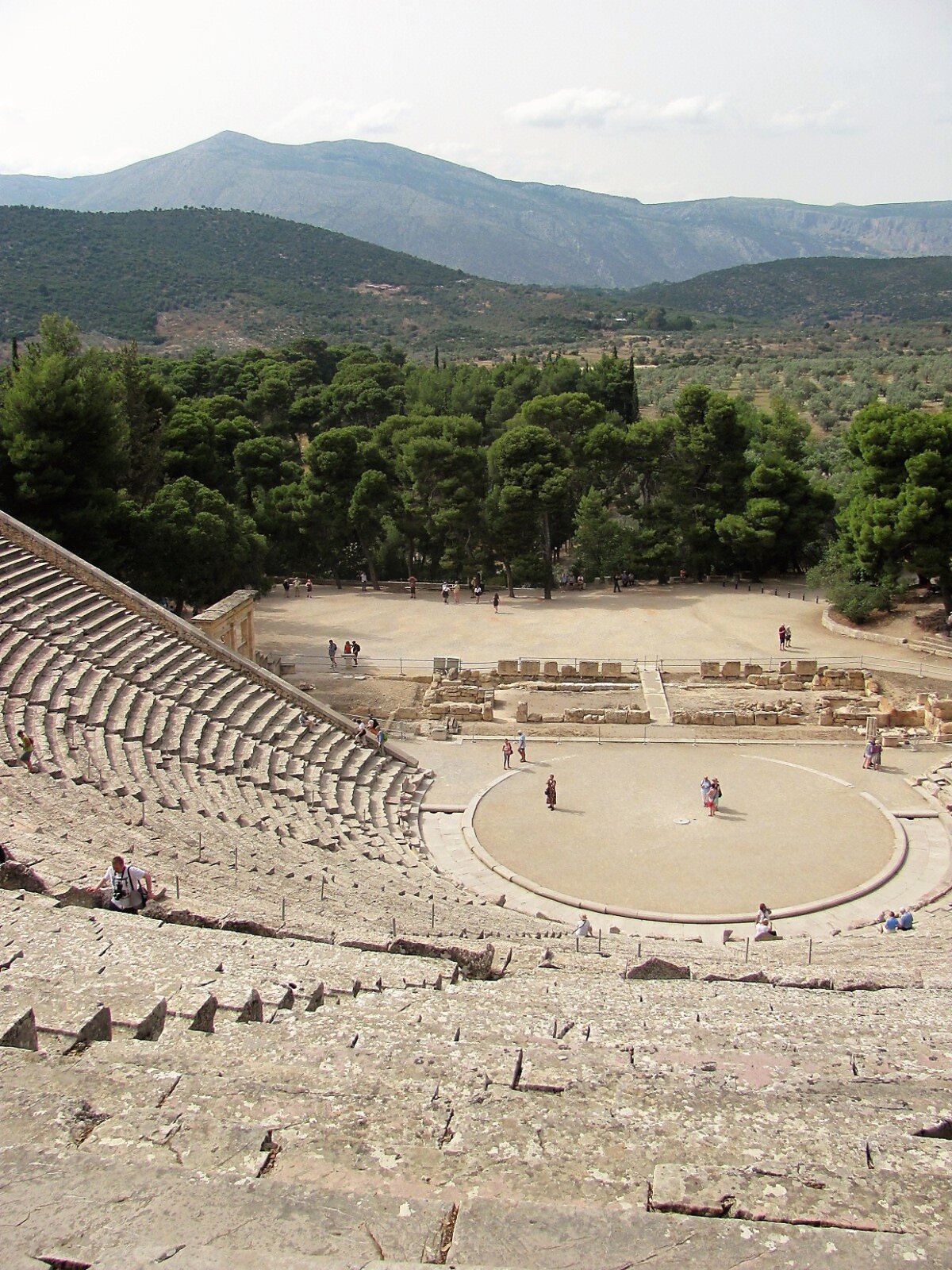 Fotografia przedstawia pozostałości starożytnego teatru w Epidauros. Zdjęcie zostało wykonane z najwyższego miejsca półkolistej schodkowej widowni. Na środku placu znajduje się okrągła orchestra – miejsce dla chóru, za nią szczątki budynków scenicznych. Za teatrem piaszczysty plac i wysokie zielone drzewa. W tyle wysokie wzgórza.