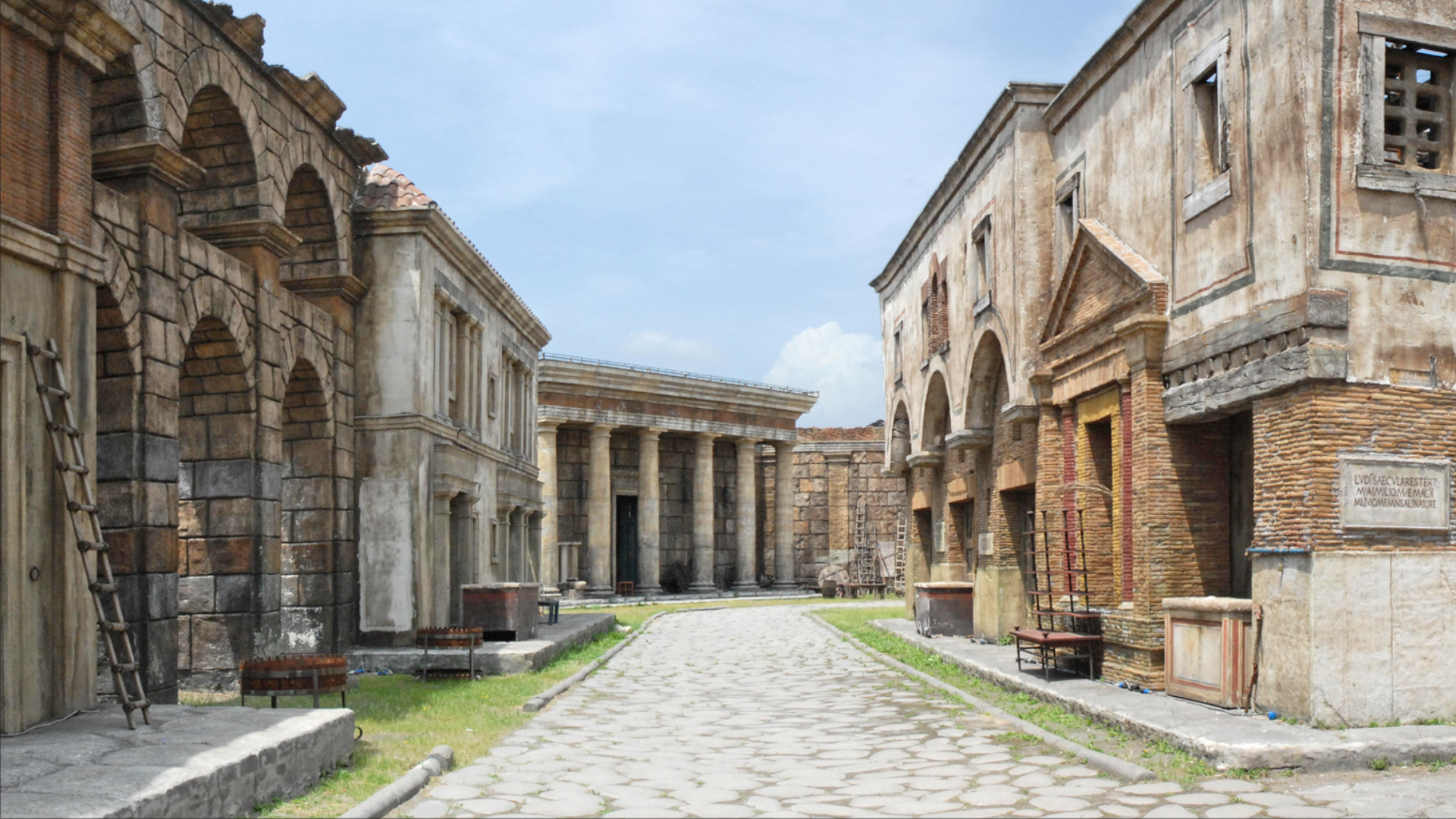 Zdjęcie przedstawia ulicę w Cinecittà, mieście kina, znajdującego się na przedmieściach Rzymu. Ulica jest kamienista. Po lewej i po prawej widoczne są budynki.