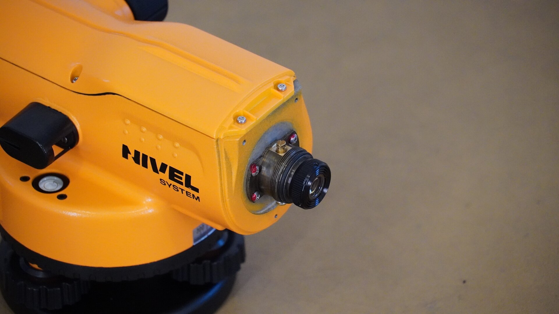 Zdjęcie ukazuje niwelator od strony okularu z perspektywy bocznej. Widoczne są śruby mocujące zespół okularu do korpusu oraz czarne pokrętła po bokach i podziałka koła poziomego poniżej.