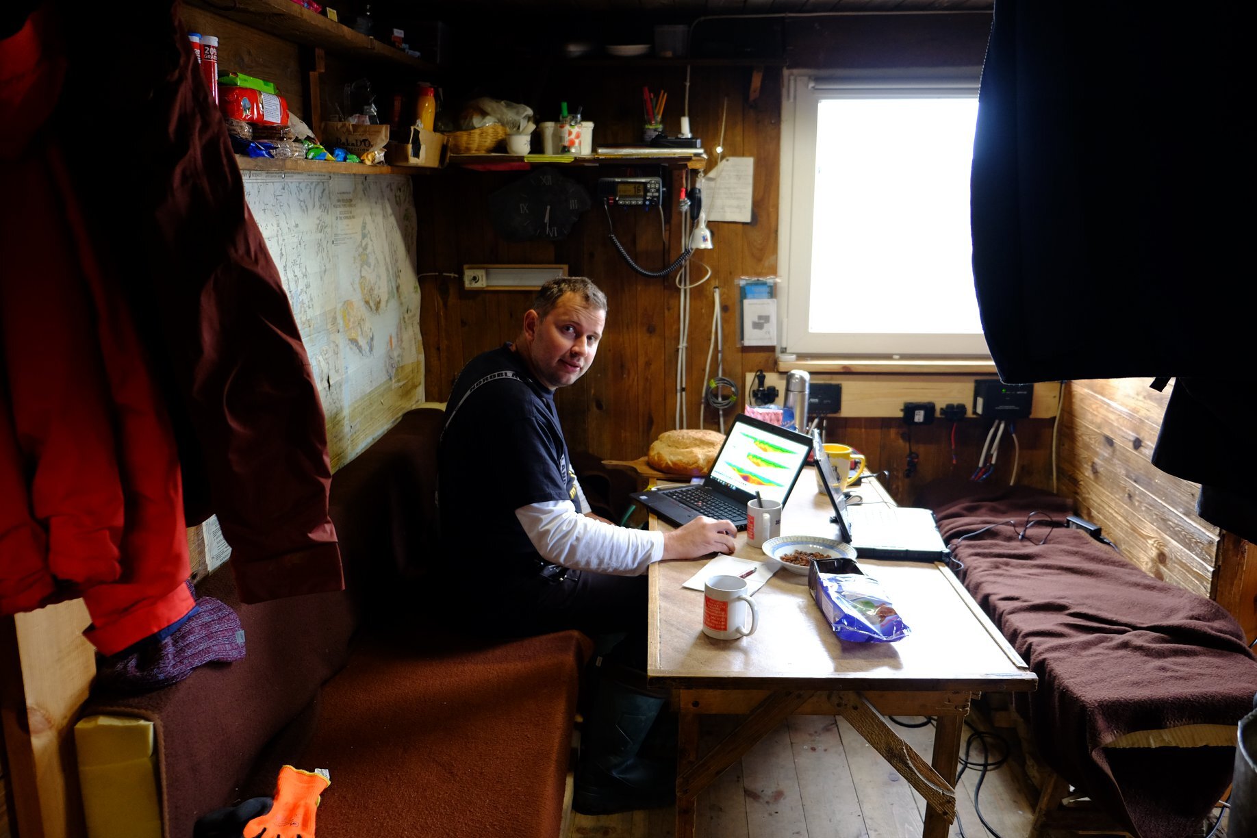 Na zdjęciu badacz wewnątrz stacji polarnej. Małe pomieszczenie, mężczyzna siedzi przy stole, na którym znajduje się komputer. Za nim mapa na ścianie, łóżka po obu stronach pomieszczenia. Drewniane ściany i podłoga, małe okno.