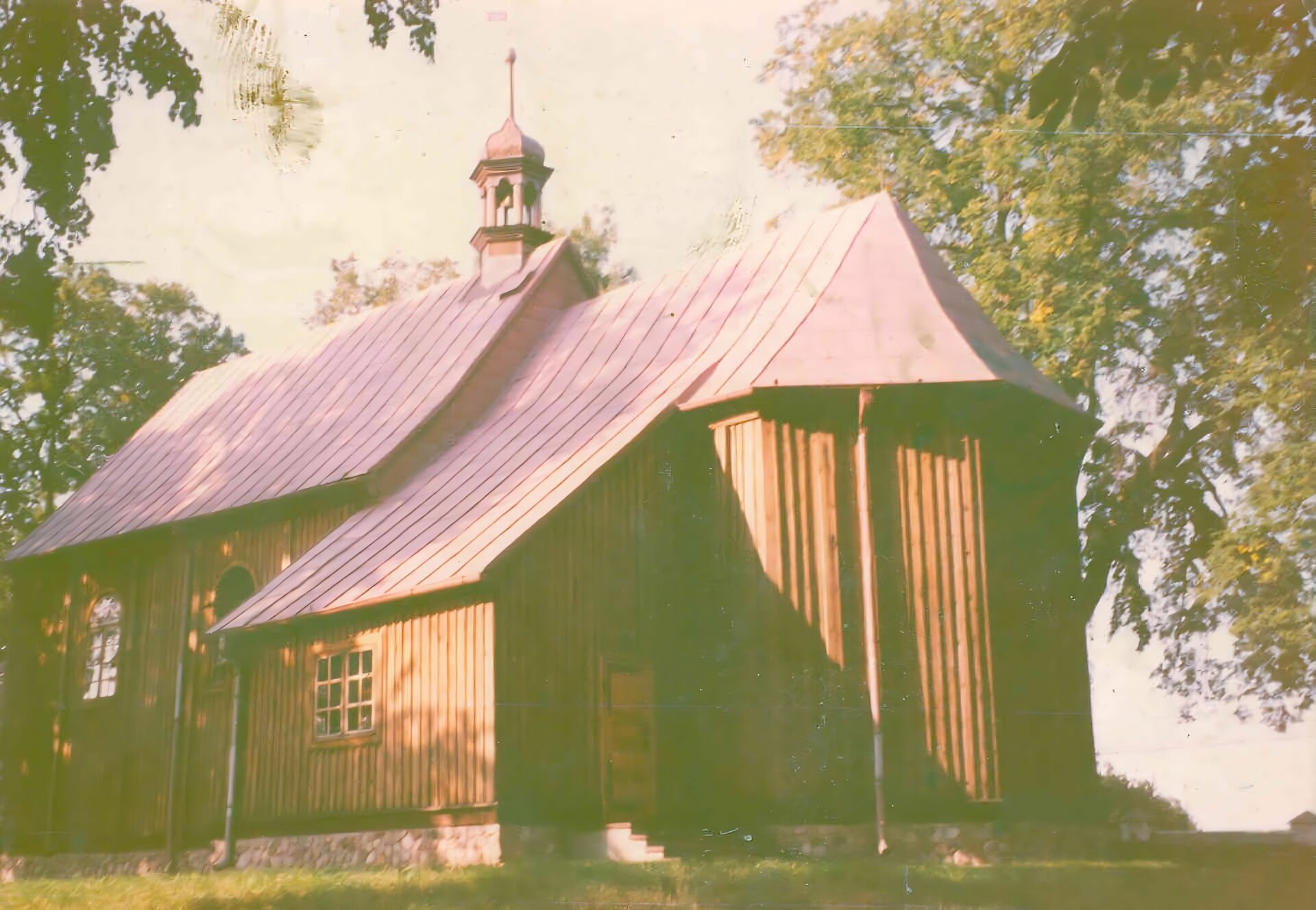 Zdjęcie przedstawia drewniany kościół od strony ołtarza. Ma on spadzisty, szary dach. Na czubku dachu jest umieszczona wieżyczka z kopułą i krzyżem. Wokół kościoła rośnie trawa oraz wysokie, zielone drzewa. Zdjęcie utrzymane jest w pastelowej kolorystyce. 