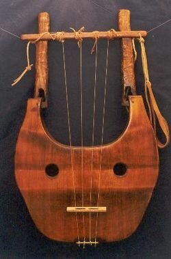 Fotografia przedstawia drewniany instrument muzyczny w kolorze brązowym. Składa się płaskiego pudła rezonansowego oraz umocowanych w nim dwóch ramion połączonych w górnej części poprzeczką. Pomiędzy korpusem a poprzeczką zostały rozciągnięte cztery struny.