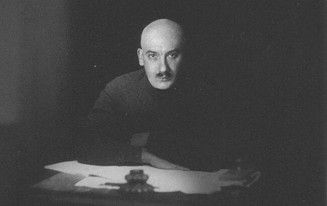 Czarno‑białe zdjęcei przedstawia Gienricha Jagodę. Siedzi on w przy biurku w ciemnym pomieszczeniu. Jest on łysy i ma krótkiego ciemnego wąsa. Na biurku leżą dokumenty. 