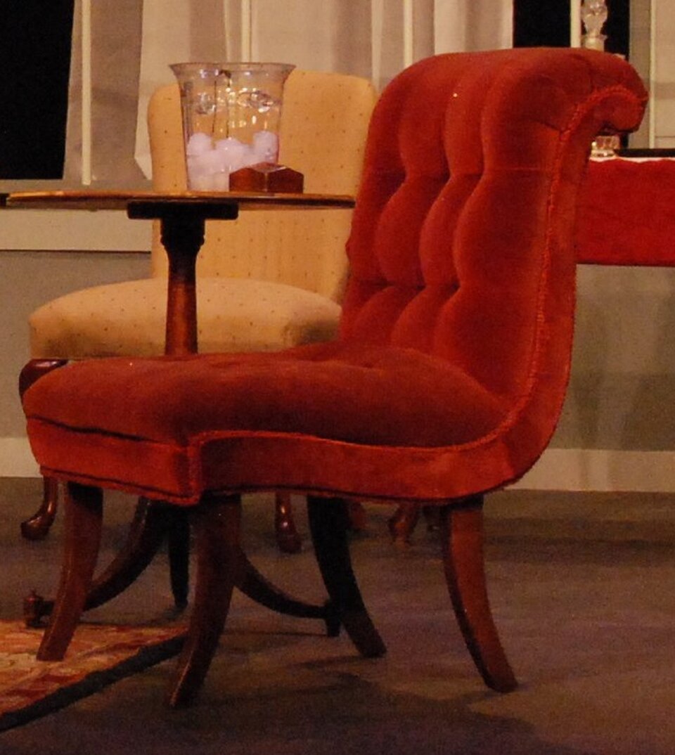Ilustracja przedstawia czerwony fotel, który jest elementem scenografii scenicznej. Mebel jest welurowy na drewnianych, wygiętych nogach. Tapicerka jest pikowana. Ustawiony jest w pomieszczeniu z okrągłym stolikiem i kremowym krzesłem.
