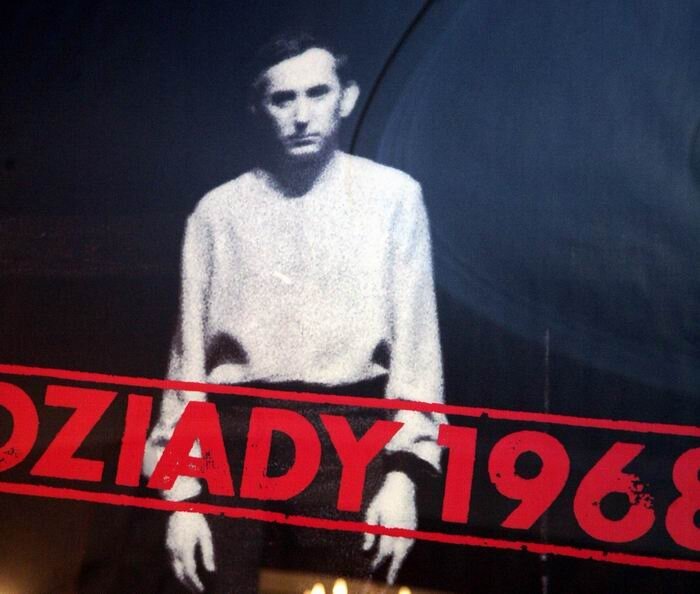 Na plakacie teatralnym znajduje się stojący mężczyzna. Ma ciemne spodnie i jasny sweter. Na nim jest czerwony napis: Dziady 1968.