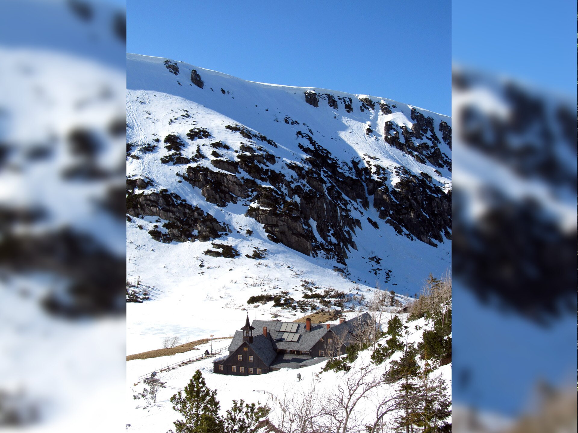 Fotografia prezentuje dolinę górską, otoczoną wysokimi ośnieżonymi szczytami. Na dnie doliny znajduje się budynek mieszkalny. Jest to schronisko górskie Samotnia w Karpaczu. Wokół schroniska nieliczne drzewa iglaste. Niebo niebieskie, bezchmurne.