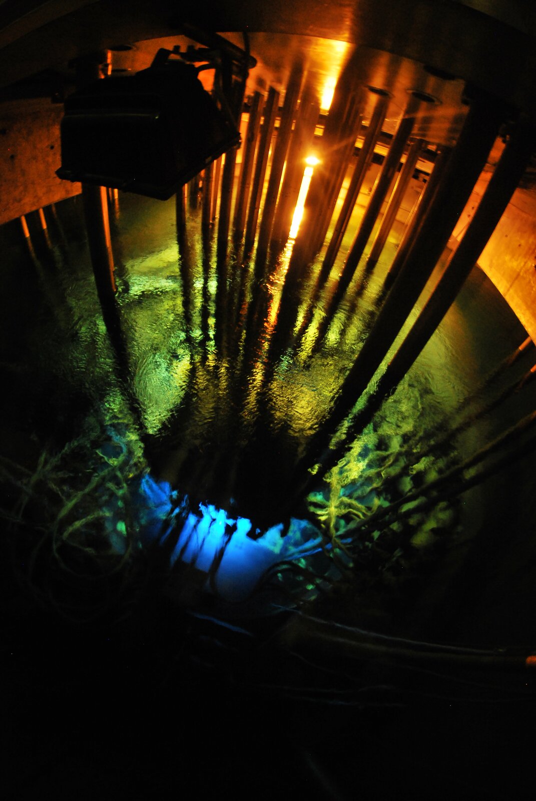 Zdjęcie przedstawia komorę reaktora w czasie jego pracy. System reaktora zanurzony jest w wodzie, spod powierzchni wydobywa się niebieskozielone światło, a znad powierzchni pomarańczowe. 