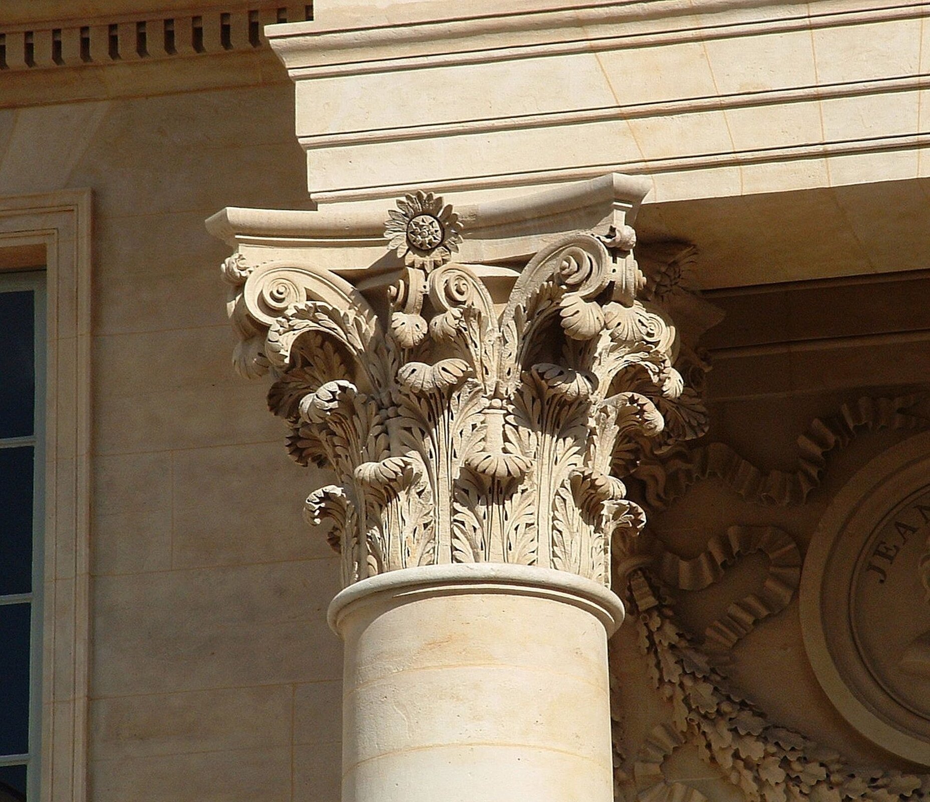 Ilustracja przedstawia zdjęcie kapitelu kolumny w stylu korynckim. Głowica dekorowana jest zwiniętymi liśćmi akantu. Nad głowicą znajduje się fragment gzymsu, a w tle widoczna jest ściana z fragmentem okna. Po prawej stronie znajduje się dekoracja z liści dębu z pomarszczoną wstęgą.