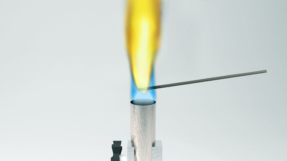 Ilustracja przedstawia metalowy pręt nad płomieniem palnika. Płomień ma kolor intensywnie żółty. 