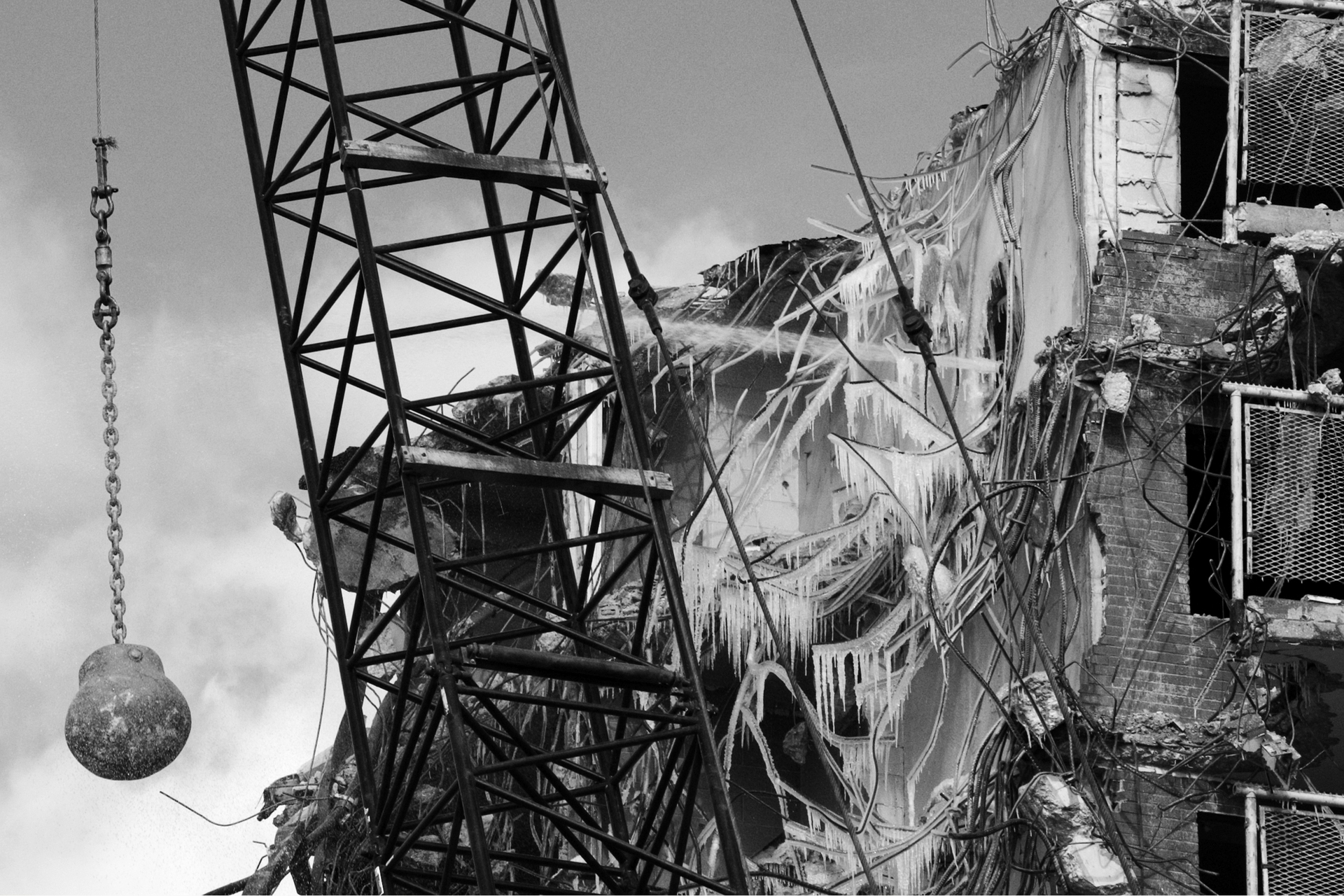 Zdjęcie przedstawia kulę burzącą zawieszoną na grubym łańcuchu na dźwigu. Widoczny fragment dźwigu w postaci ażurowej metalowej konstrukcji. W tle widać ruiny budynku, strzępy zwisających materiałów. Fotografia jest czarnobiała.