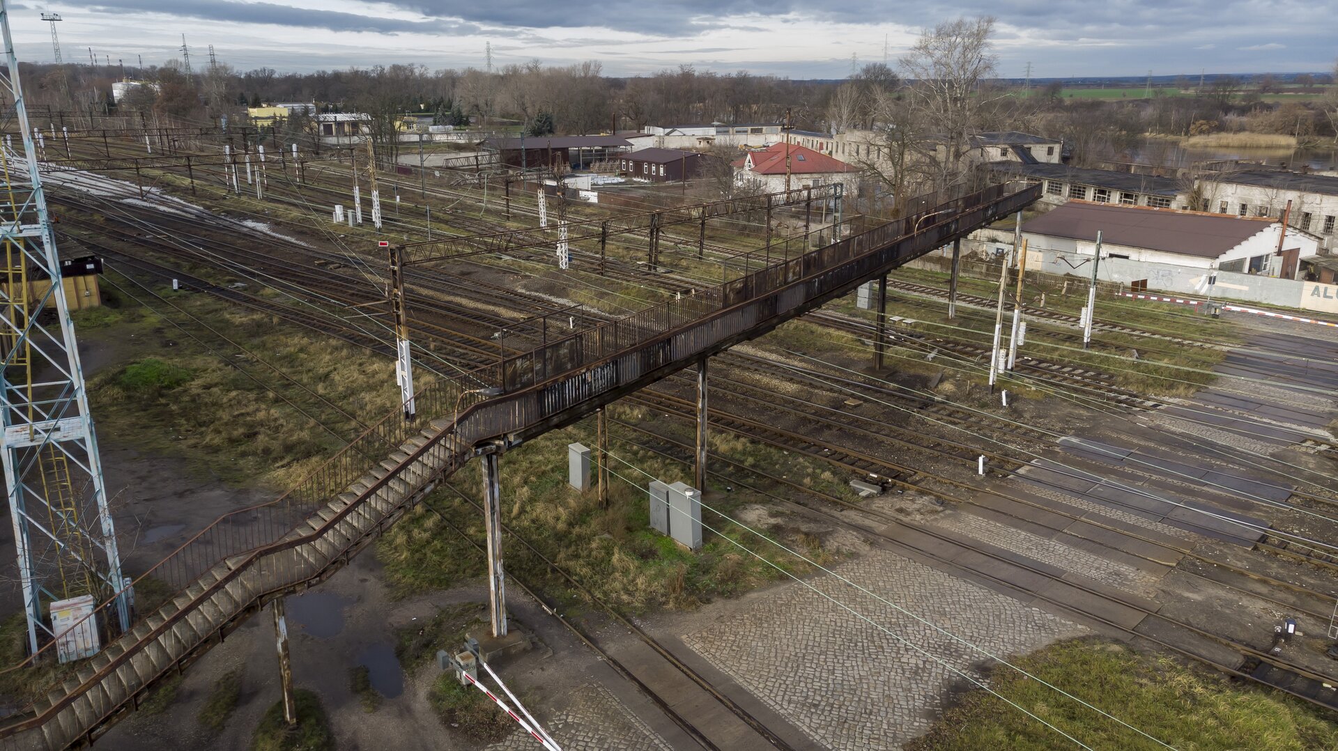 Ilustracja przedstawia widok z góry na kładkę dla pieszych, która znajduje się nad torowiskiem składającym się z dziesięciu torów. Kładka jest wykonana z metalu, a do niej prowadzą schody. Na niektórych elementach kładki widoczna jest rdza. Szlabany na przejeździe kolejowym są zamknięte. Po lewej stronie od kładki znajduje się wysoki przekaźnik, a w tle widać budynki oraz chmury. Kładka umożliwia pieszym bezpieczne przekroczenie torów kolejowych.