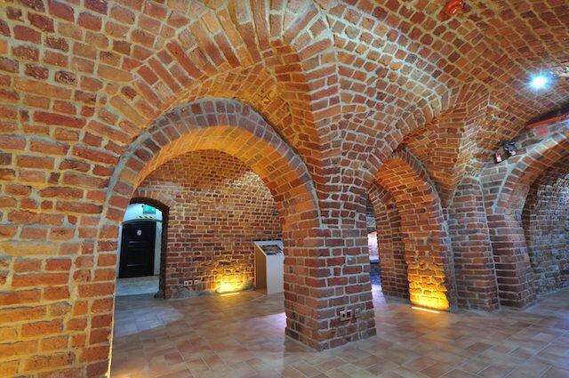 Zdjęcie przedstawia piwnice. Widoczne są łukowate sklepienia zbudowane z czerwonych cegieł.