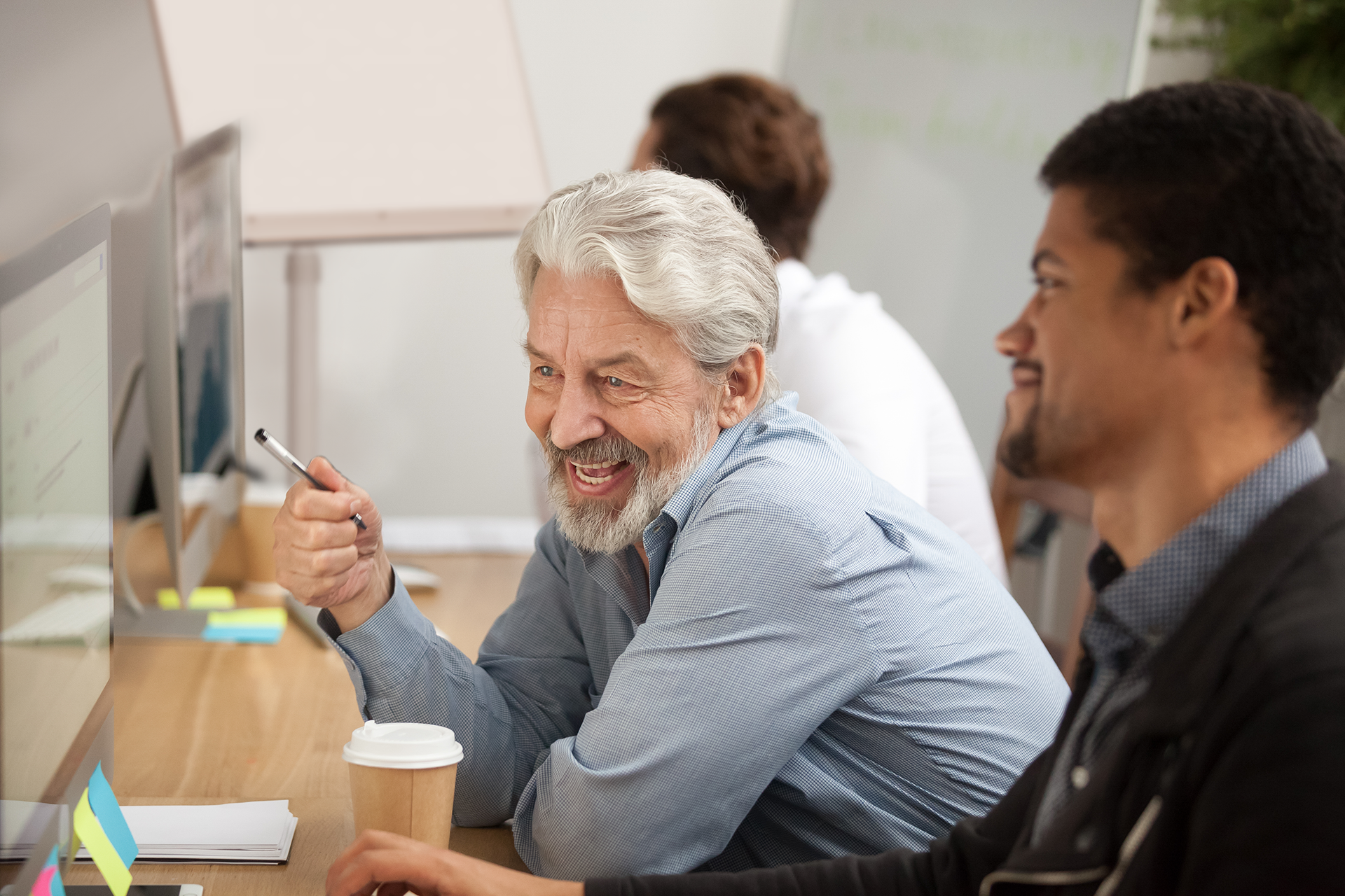Zdjęcie przedstawia dwóch mężczyzn siedzących przy biurku i patrzących w monitory. Mężczyzna w centralnej części fotografii ma siwe włosy i brodę. W dłoni trzyma długopis i patrzy na komputer. Obok niego, po prawej stronie zdjęcia siedzi mężczyzna o ciemnej karnacji, z krótką brodą i wąsami. W prawej dłoni trzyma myszkę od komputera. W tle widać kolejnego mężczyznę.