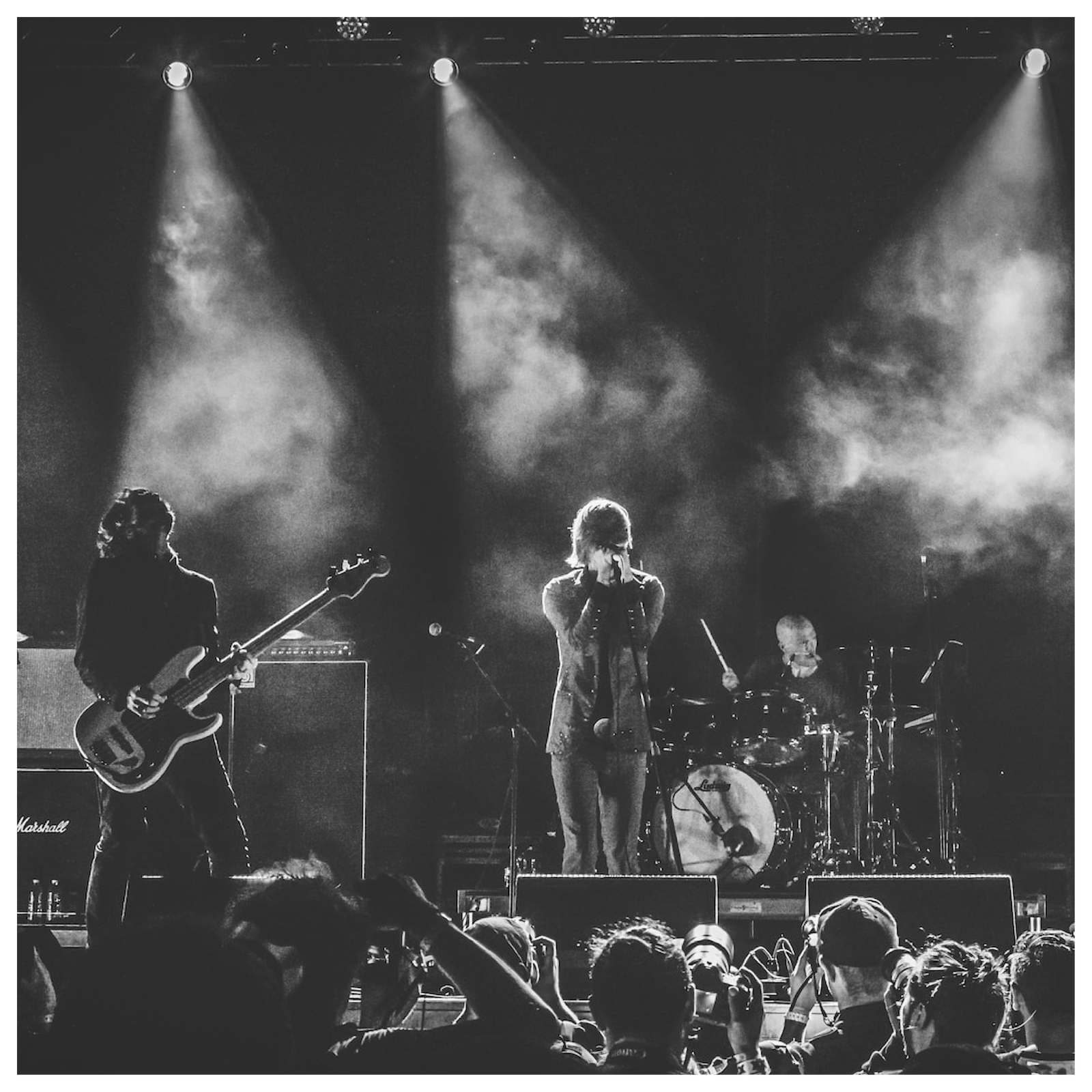 Zdjęcie jest czarno‑białe i przedstawia koncert. Na scenie widoczny jest wokalista przed mikrofonem, gitarzysta i perkusista. Pod sceną rzesza fanów.