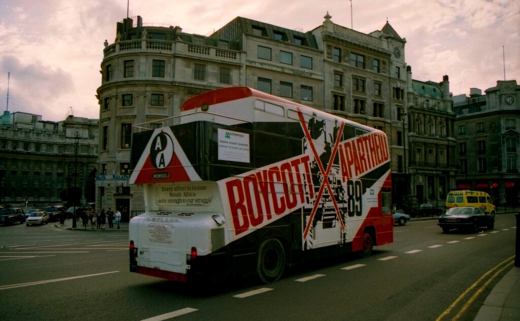 Zdjęcie przedstawia fragment miasta. Ulicą jedzie piętrowy autobus wymalowany wzdłuż jednego z boków pochyły napis po angielsku: bojkot apartheidu. W tle zabudowania i ulice, na których jadą samochody. Po chodnikach idą ludzie. 