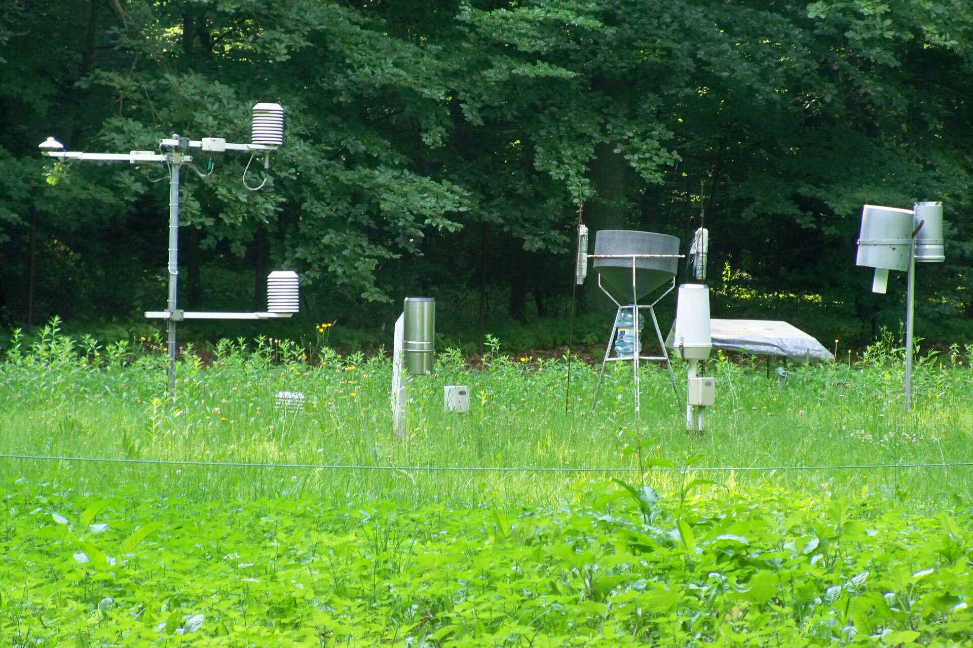 Zdjęcie przedstawia ogródek meteorologiczny. Ogródek otoczony jest płotkiem z siatki. Wewnątrz ogródka znajdują się przyrządy meteorologiczne, w tym klatka meteorologiczna w kształcie drewnianego pudełka. Wewnątrz klatki znajdują się przyrządy pomiarowe. Na terenie ogródka widoczne są wiatromierz na wysokim słupie oraz deszczomierz.