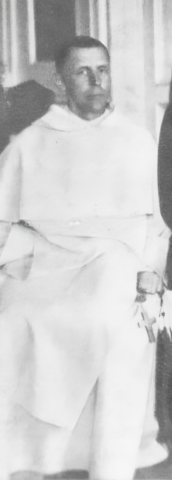 Przedstawia zdjęcie młodego mężczyzny w habicie dominikańskim, który prawą dłoń schował za szkaplerz, a w lewej dłoni trzyma różaniec z dużym krzyżem. Jest bez zarostu, krótko ostrzyżony. Na dalszym planie zdjęcia widać wnętrze kościoła. 