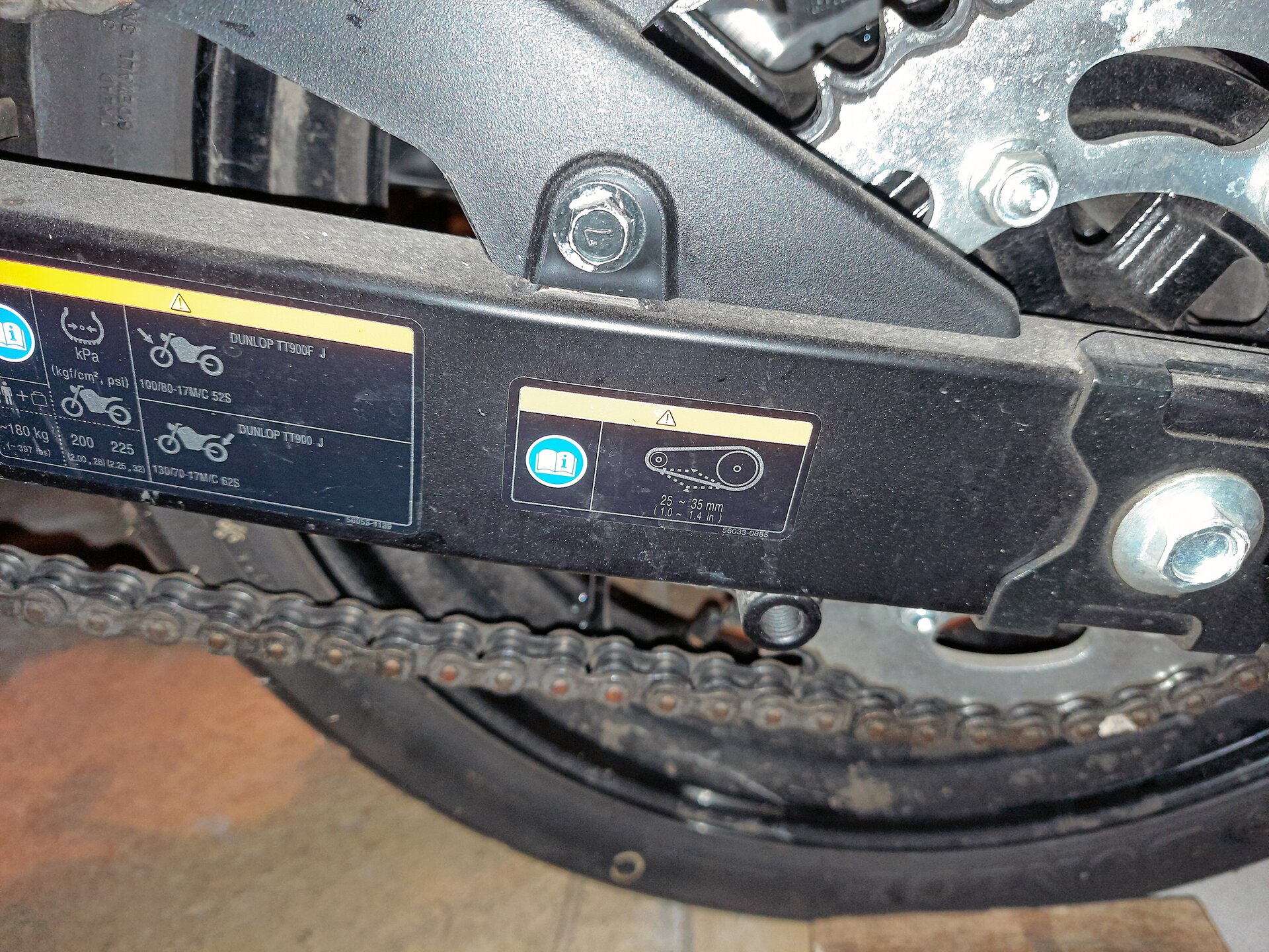 Na ekranie zbliżenie na naklejkę informującą o właściwym luzie łańcucha napędowego. Jest umieszczona na wahaczu tylnym.