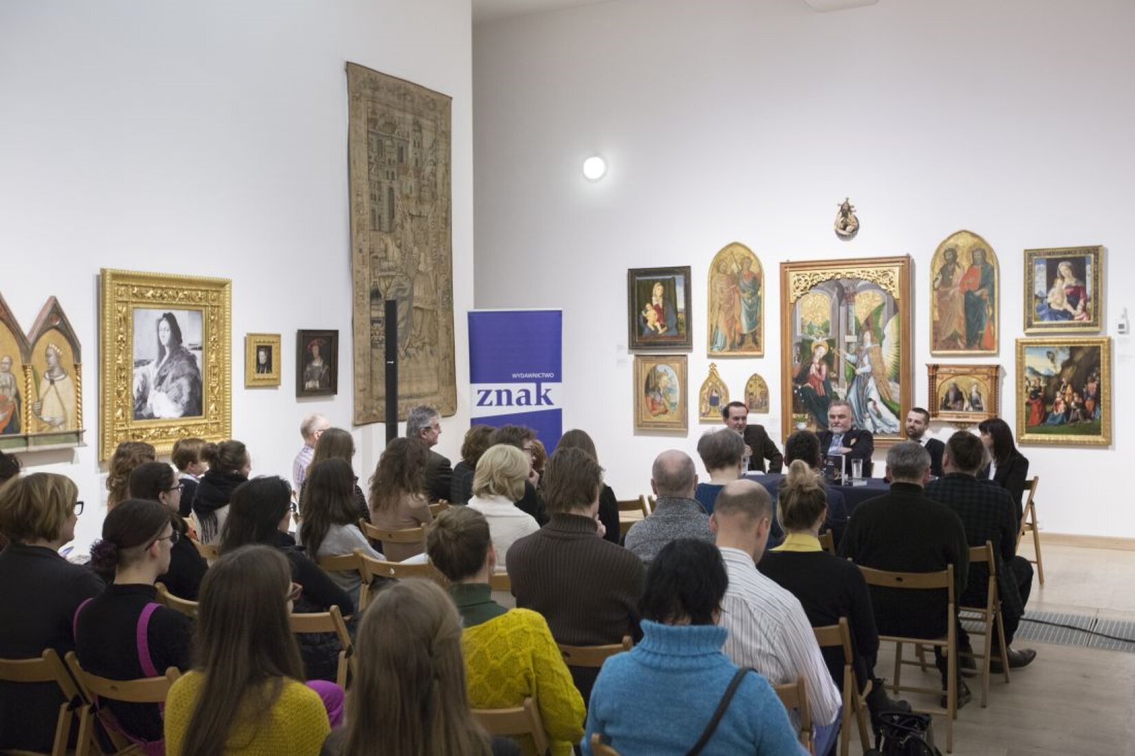 Na fotografii ukazano pomieszczenie, w którym znajduje się wiele ludzi. Siedzą na drewnianych krzesłach, tyłem do obserwatora. Zwróceni są do trzech mężczyzn, siedzących w centrum. Przed mężczyznami stoi stół, na którym znajdują się książki oraz szklanka wody. Na białych ścianach pomieszczenia znajdują się liczne obrazy. W rogu pomieszczenia, znajduje się biało‑niebieski baner z napisem „Wydawnictwo znak”.