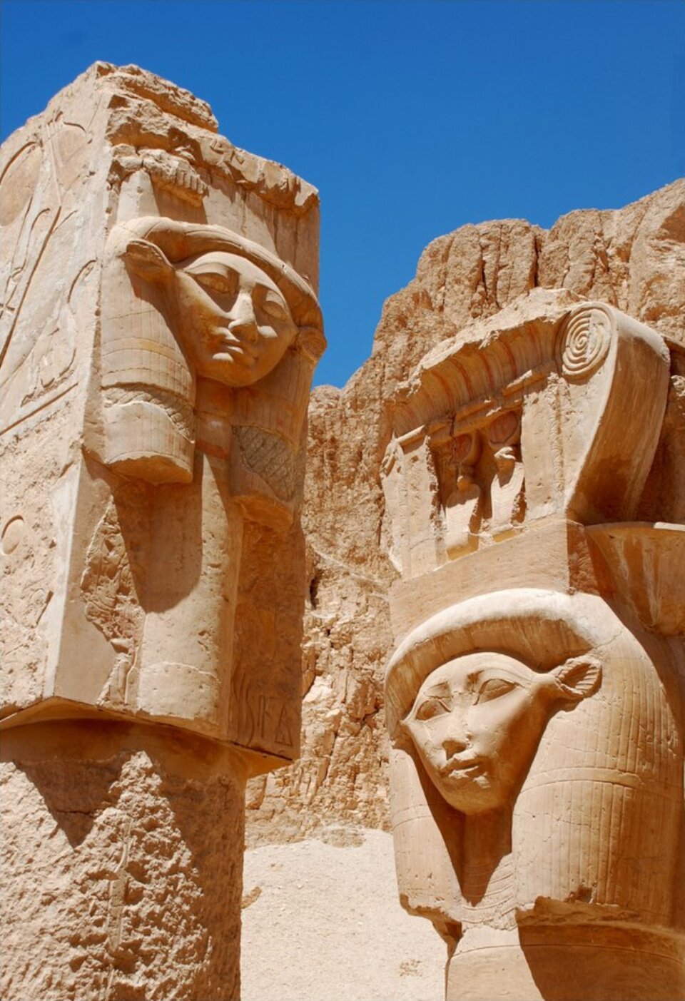 Fotografia przedstawia kolumnę w stylu hatoryckim stojącą nieopodal sfinksa. Kolumna składa się z trzonu oraz głowicy. Głowica kolumny to płaska powierzchnia, na której umieszczona została rzeźba głowy postaci o szerokim rozstawie oczu i neutralnym wyrazie twarzy. Na głowie postaci jest nakrycie głowy - korona faraona. Na bocznej ścianie głowicy nieczytelne reliefy.