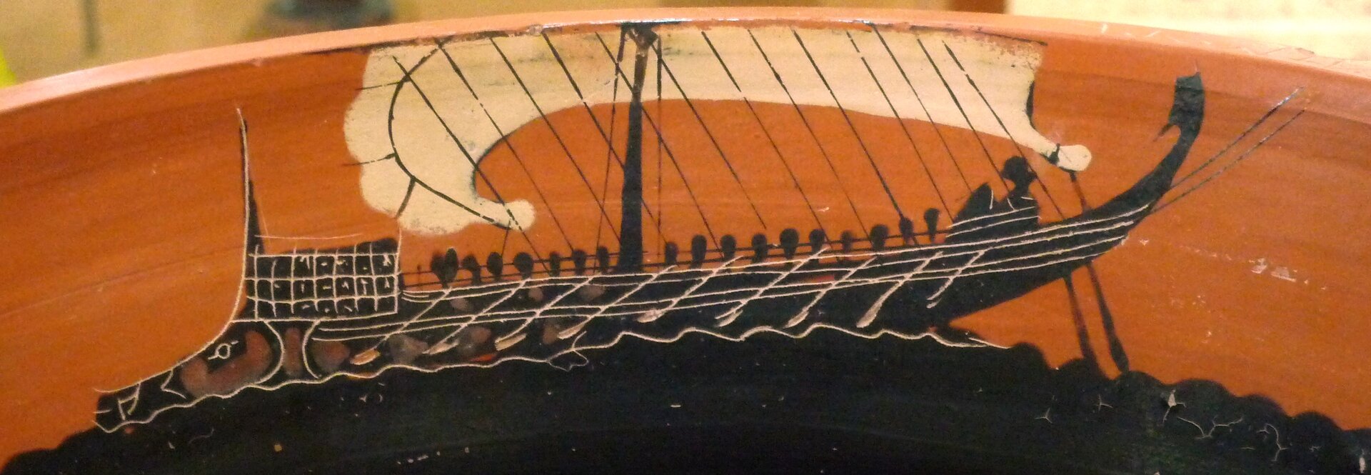 Ilustracja przedstawia fragment naczynia ozdobionego rysunkiem antycznego okrętu morskiego. Dziób oraz przednia część kadłuba są koloru czarnego, zaś rufa i tylna część okrętu są ciemnoczerwone. Na pokładzie siedzą ciemne postacie trzymające wiosła, nad nimi kremowy żagiel. Statek płynie po wodzie, która jest czarna. Tło stanowi pomarańczowe niebo. Dzieło jest przykładem malarstwa czarnofigurowego.