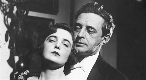 Zdjęcie przedstawia mężczyznę i kobietę. Kobieta ma półprzymknięte oczy i opiera głowę na ramieniu mężczyzny, stojącego za nią. Kobieta ma krótkie, kręcone włosy. Mężczyzna wpatruje się w kobietę. Ubrany jest w białą koszulę i marynarkę. Ma lekko kręcone włosy zaczesane do tyłu. 