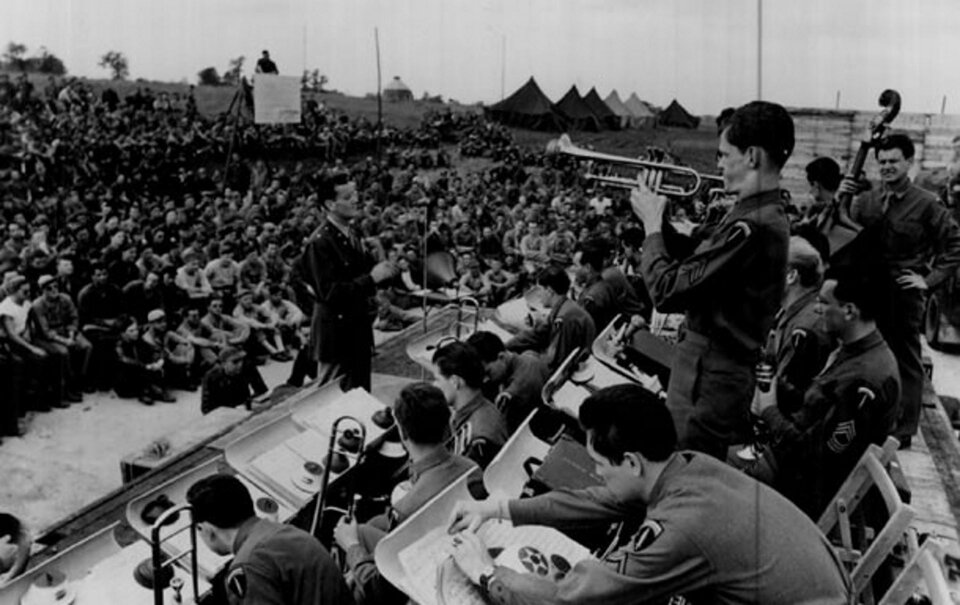 Ilustracja przedstawia Glenn Miller z orkiestrą wojskową. Przed orkiestrą stoi mężczyzna - dyrygent. Jeden z żołnierzy stoi i gra na instrumencie muzycznym. W oddali widać tłumy słuchaczy.