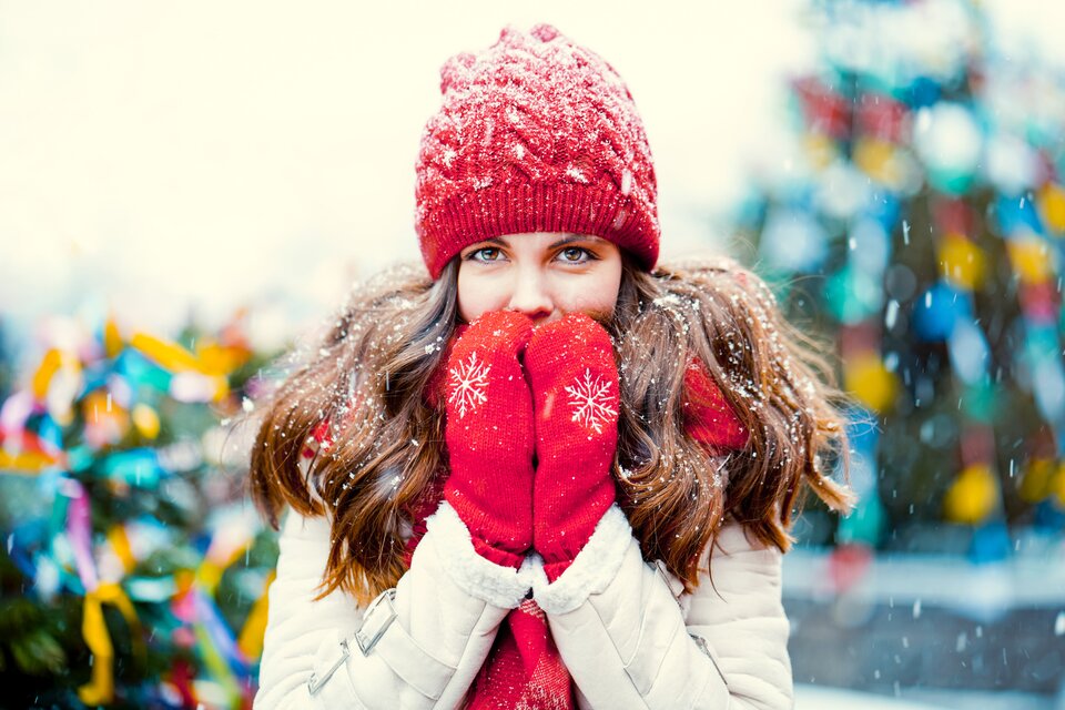Zdjęcie przedstawia dziewczynę w czerwonej czapce i rękawiczkach z płatkami śniegu na ubraniu; w tle świąteczne ozdoby i padający śnieg.