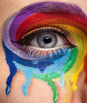 Ilustracja przedstawia fotografię z wykadrowaną częścią twarzy z prawym okiem. Prawie cały kadr jest pokryty farbami w intensywnych kolorach, które ściekają po policzku. 