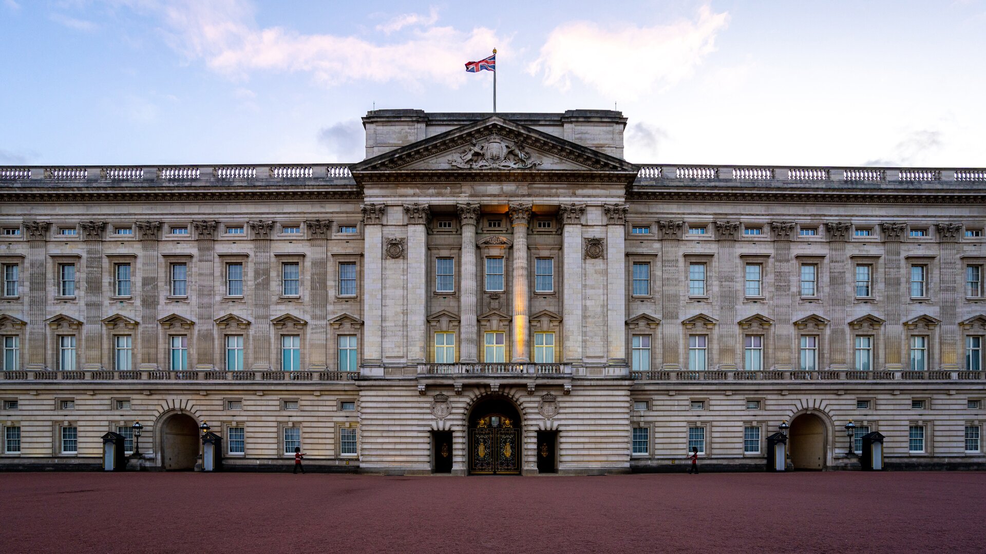 Zdjęcie przedstawia fasadę pałacu  Buckingham . Budynek jest szary, dwukondygnacyjny, z wysokimi oknami i betonowymi barierkami na dachu. Wejście jest stylizowane na antyczne, posiada sześć kolumn zwieńczonych panteonem.