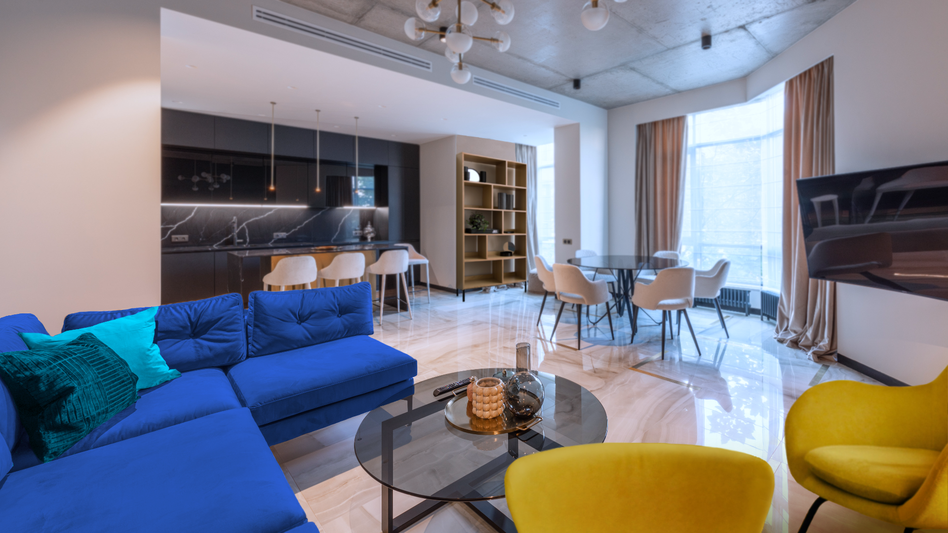 Zdjęcie przedstawia obszerny salon i kuchnię. W salonie i kuchni znajduje się wiele mebli. Po lewej stronie znajduje się duża kanapa narożna. Przed kanapą stoi okrągły stolik kawowy.  Po prawej, obok stolika stoją dwa fotele.  Na ścianie po prawej wisi telewizor. Na drugim planie, po prawej pod oknem stoi stół z krzesłami. Za stołem po prawej znajduje się duże okno, a po lewej od okna stoi regał. Po lewej stronie od regału znajduje się aneks kuchenny. 