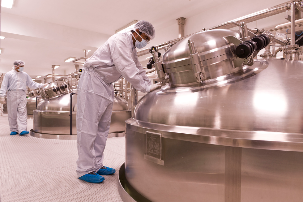 Fotografia przedstawia wnętrze zakładu, produkującego insulinę przy pomocy bakterii. Stoją olbrzymie metalowe kadzie, w których hoduje się odpowiednio zmodyfikowane mikroorganizmy. Przy kadziach ludzie w białych kombinezonach, ochraniaczach na buty, rękawiczkach i maseczkach.
