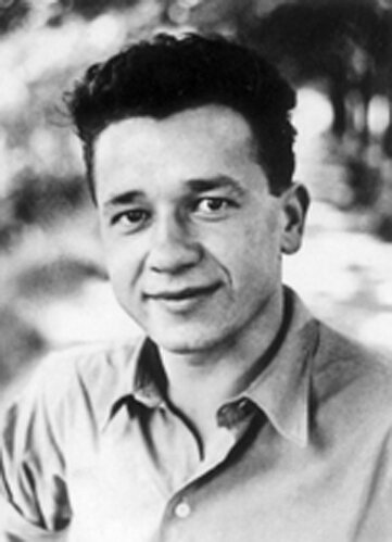 Czarno‑białe zdjęcie przedstawia portret mężczyzny. Ma podłużną twarz, ciemne, zaczesane ku górze włosy oraz szeroki nos. Mężczyzna jest uśmiechnięty. Ubrany jest w jasną koszulę.