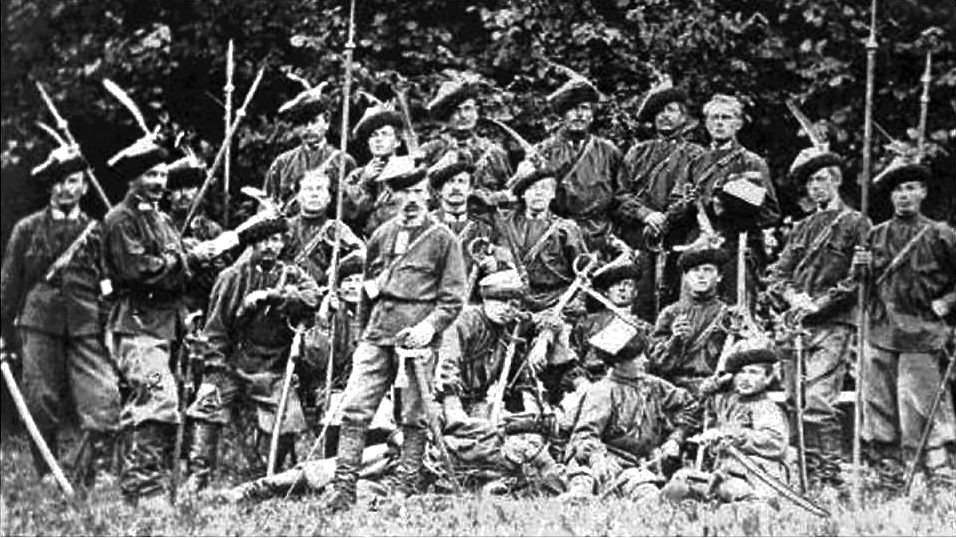 Zdjęcie przedstawia grupę mężczyzn stojących i siedzących na polanie i pozujących do zdjęcia. Mężczyźni mają na głowach rogatywki, w dłoniach trzymają kosy z ostrzami postawionymi na sztorc.  