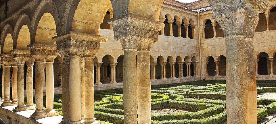 Fotografia przedstawia dziedziniec przed którym stoi kolumnada podtrzymująca loggię budynku. Budynek, jak i kolumny są zbudowane z kamienia, ktory ma kolor złotego piasku. Głowice na kolumnach mają kształt ściętego ostrosłupa, którego ściany zostały bogato przyozdobione wymodelowanymi elementami w kształcie liści akantu. Głowice opierają się na dwóch lub na czterech kolumnach. Na dziedzińcu rosną żywopłoty, które tworzą labirynt.