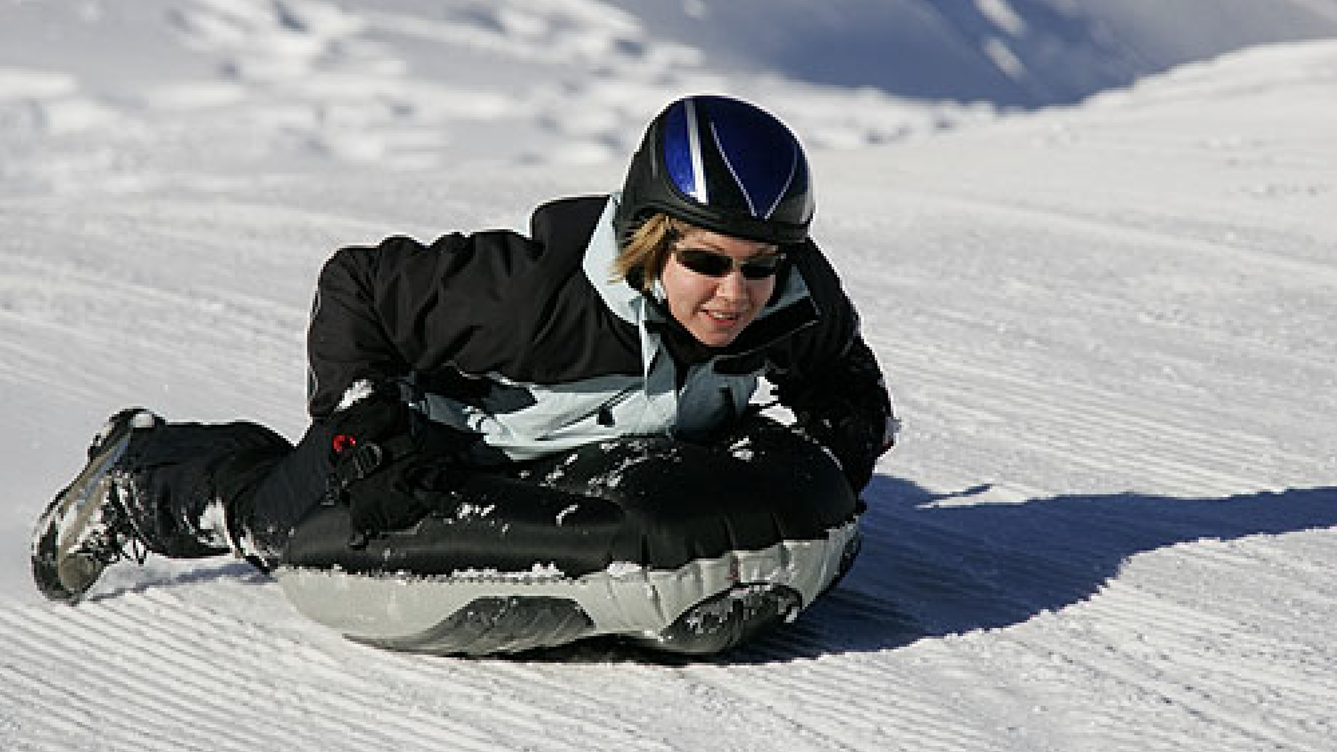 Zdjęcie przedstawia kobietę w kombinezonie narciarskim, kasku i ciemnych okularach. Kobieta leży na brzuchu na powietrznej desce i zjeżdża po śniegu.