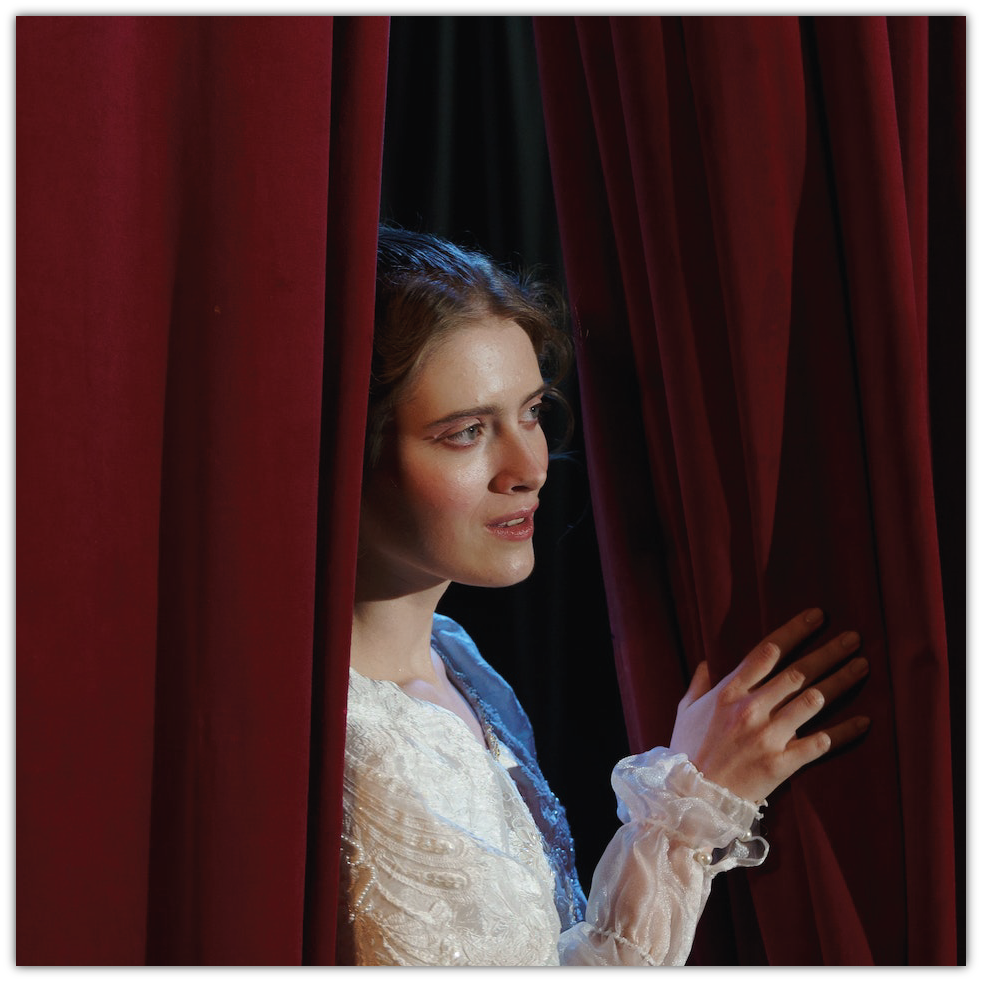 Zdjęcie przedstawia kobietę w białej sukni spoglądającą przez czerwoną, teatralną kurtynę.