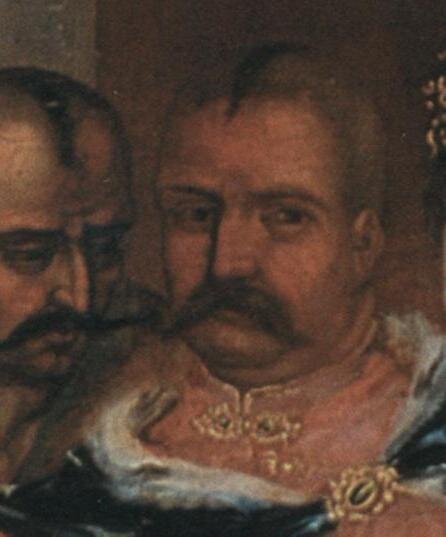 Obraz przedstawia Iwana Sulyme. Jest do duży mężczyzna. Ma długiego ciemnego wąsa i jest łysy. Na głowie ma pozostawiony jedynie kozacki kosmyk. Jest ubrany w pomarańczowe szaty. Po jego lewej stronie stoi inny kozak szepcze coś do Iwana.