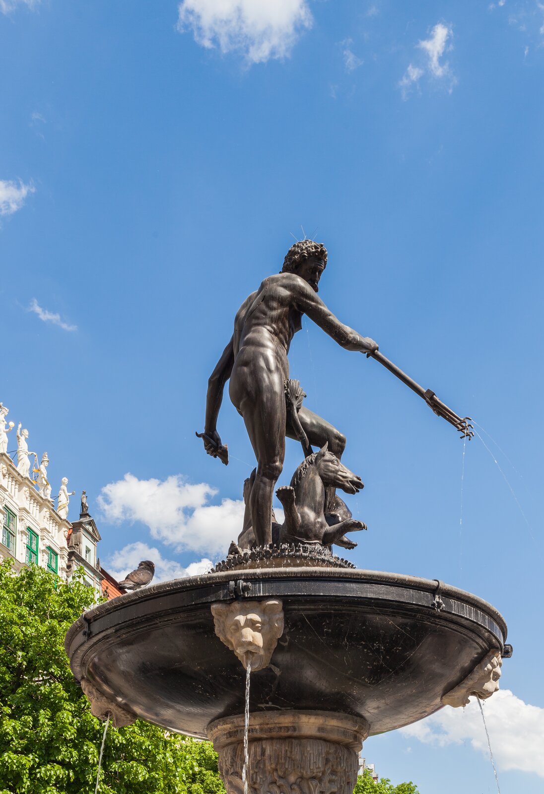 Zdjęcie przedstawia fragment fontanny z rzeźbą Neptuna, czyli rzymskiego boga wód, chmur i deszczu. Jest to mężczyzna trzymający w ręku charakterystyczny trójząb, czyli długi kij zakończony trzema ostrzami. W tle widać fragment kamienicy, zielone drzewo i błękitne niebo z jasnymi chmurami.