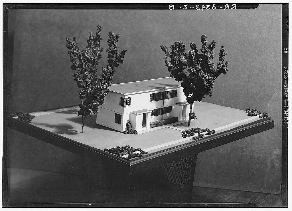 Na zdjęciu jest makieta- projekt domu. Kształt domu przypomina prostopadłościan. Dom ma okna i dwoje drzwi. Przy domu stoją dwa drzewa. 