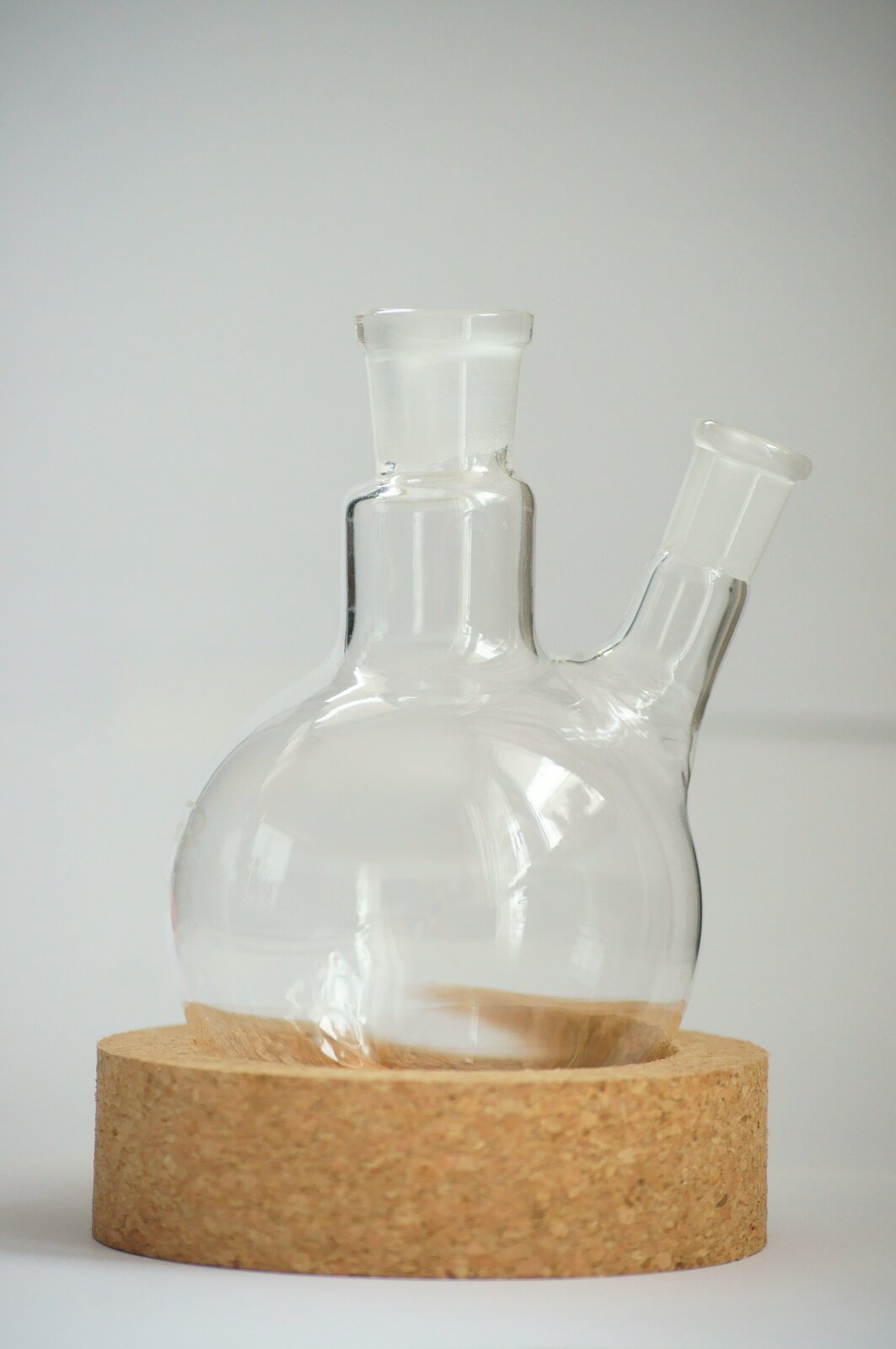 Na zdjęciu przedstawione jest kolba okrągłodenna dwuszyjna ze szlifami. Kolba stoi w guku, czyli podstawce, w tym przypadku wykonanej z korka.