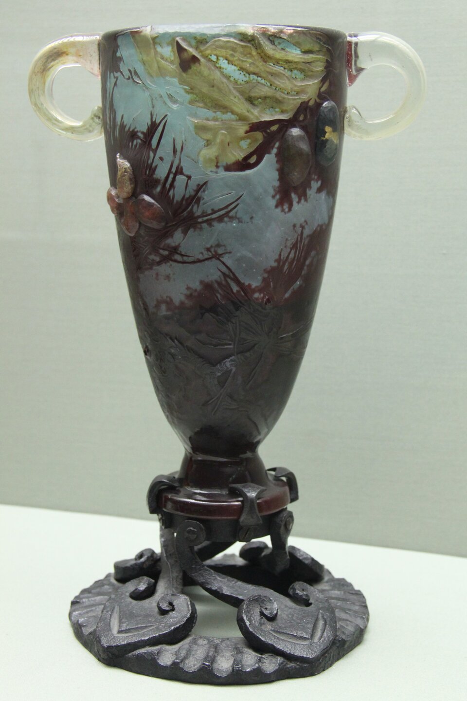 Ilustracja przedstawia kielichowate naczynie wykonane ze szkła, które jest osadzone na żelaznej, czarnej podstawce. Naczynie ma wyrzeźbione gałązki w kolorze brązowym. Wśród brązowych traw siedzi motyl żółto‑czerwony. W rogu naczynia znajduje się zielona gałązka z owocami papai. Dwa ucha są przezroczyste.