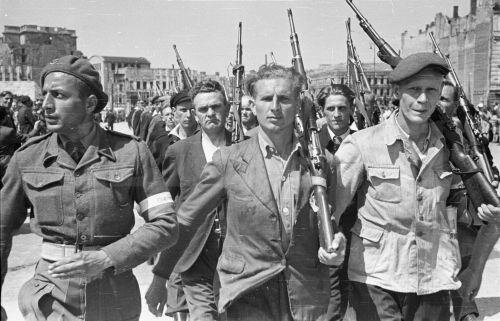 Czarno‑białe zdjęcie przedstawia maszerujących członków Ochotniczej Rezerwy Milicji Obywatelskiej. Są oni ubrani z codzienne stroje. Każdy z nich ma na ramieniu karabin. Po lewej stronie maszeruje wojskowy w mundurze, czapce i biało czerwoną przepaską na ramieniu. W tle widoczne są liczne budynki. 