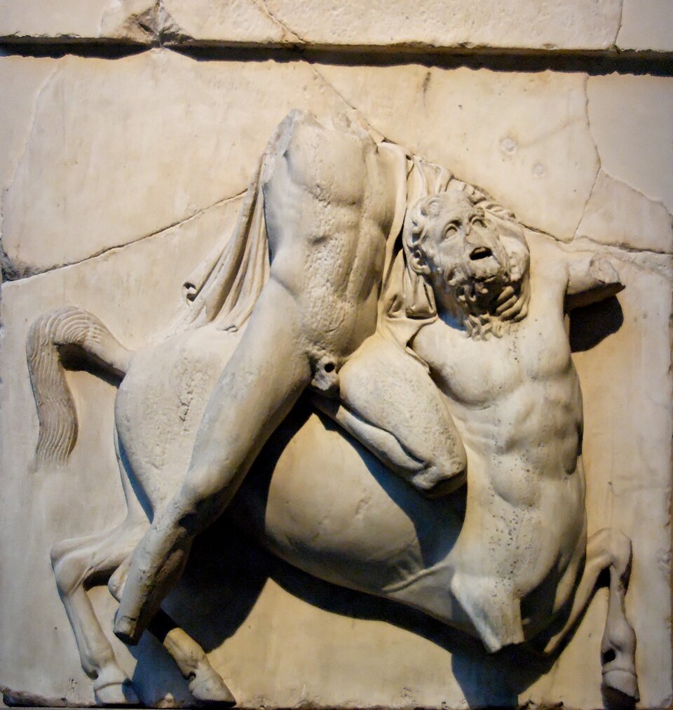 Ilustracja przedstawia Lapitę walczącego z centaurem, metopę południową II z Partenonu w Atenach. Ukazana jest dynamiczna scena walki wyrzeźbiona w marmurze: centaur wspina się na tylne nogi i pochyla ku przeciwnikowi, obejmując go ramieniem, podczas gdy Lapita odchyla się do tyłu w gwałtownym geście obrony. Obie postacie są częściowo uszkodzone - brakuje głów i fragmentów kończyn, lecz zachowane partie ukazują silne napięcie mięśni i głębokie podcięcia charakterystyczne dla wysokiego reliefu. Tło stanowi gładka płyta metopy.