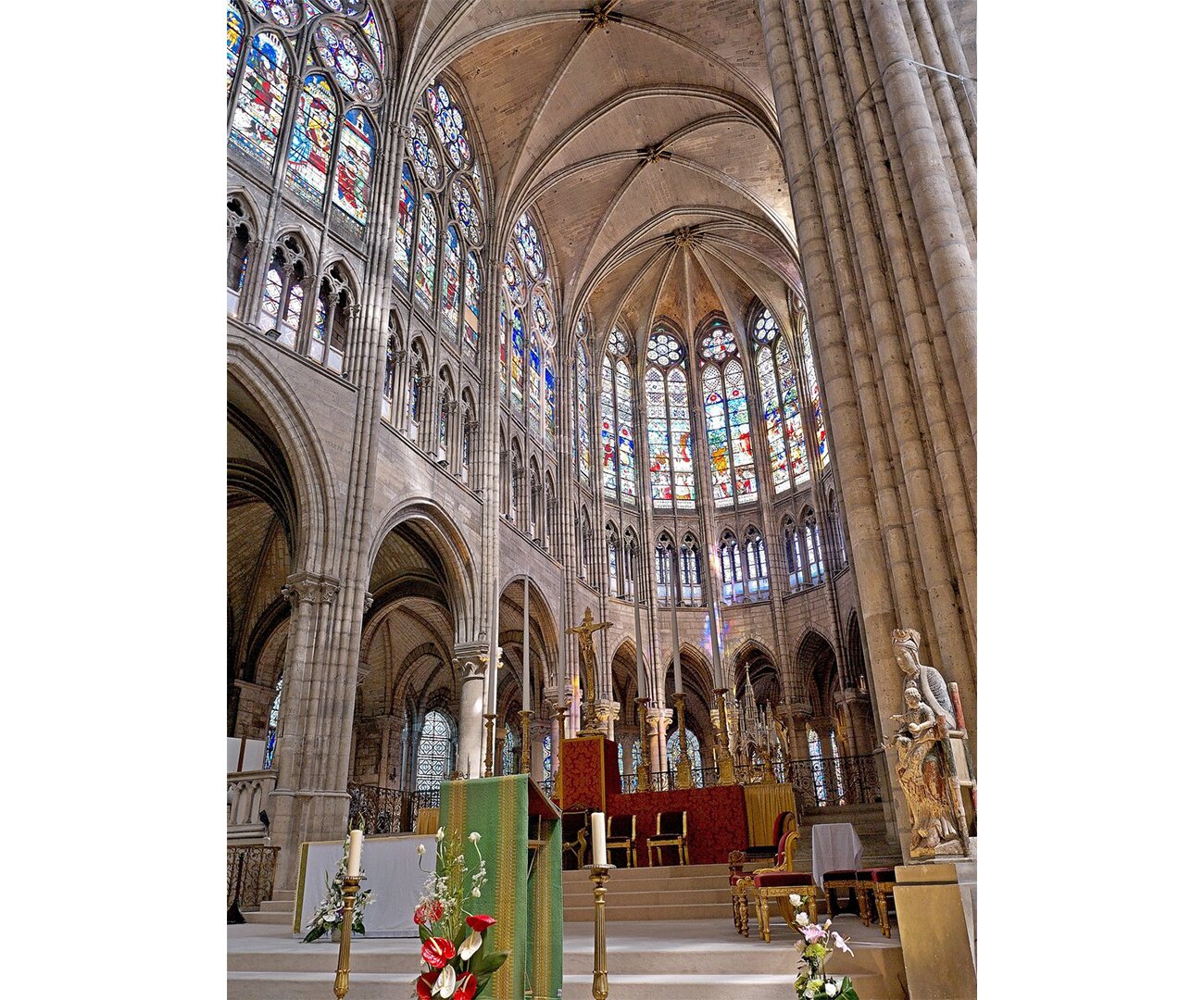 Fotografia barwna przedstawiająca Prezbiterium nawy głównej kościoła św. Denisa, XIII wiek. Uwidocznione zostały wewnętrzne sklepienia, wyżłobione elementy jak i duży witraż składający się z małych, barwnych okienek szklanych.