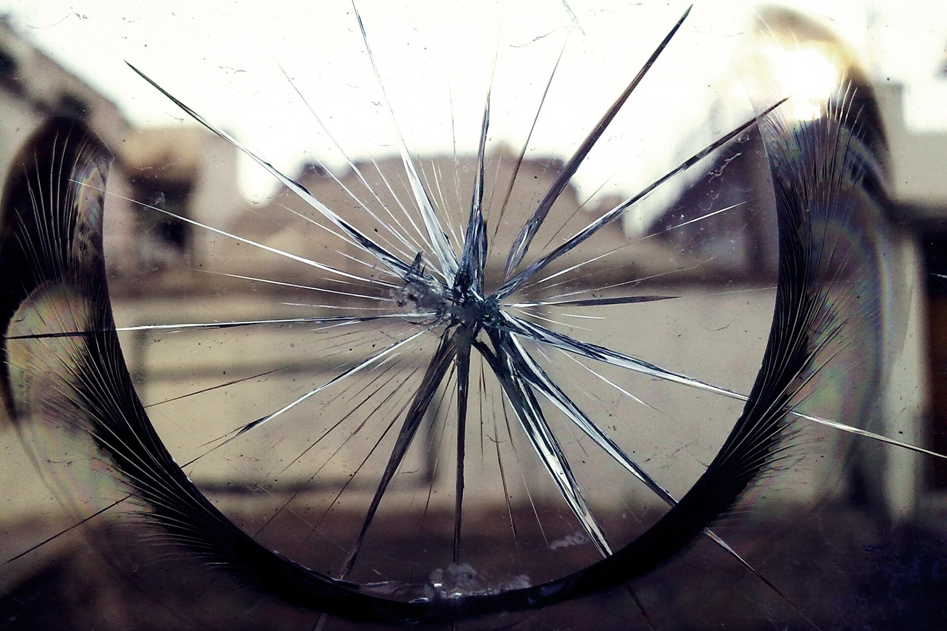 Fotografia przedstawia pęknięcie na szklanej powierzchni, ukazane z bliska. Pęknięcie ma kształt gwiazdy. Za szklaną powierzchnią w tle nieostry widok na zabudowania.